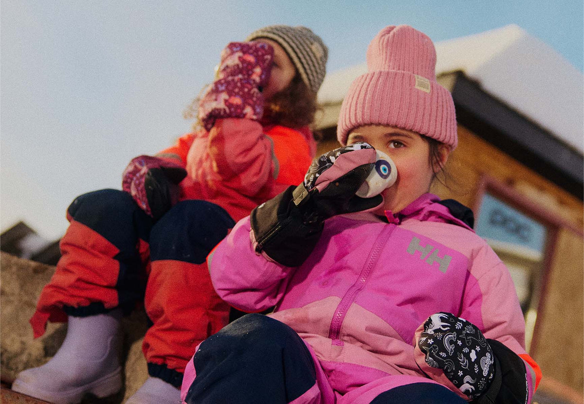 Two children in winter ski gear sitting outdoors. The child in the foreground wears a pink jacket, pink beanie, black gloves, and dark pants, while the child behind wears an orange jacket, patterned pink gloves, and a striped beanie.