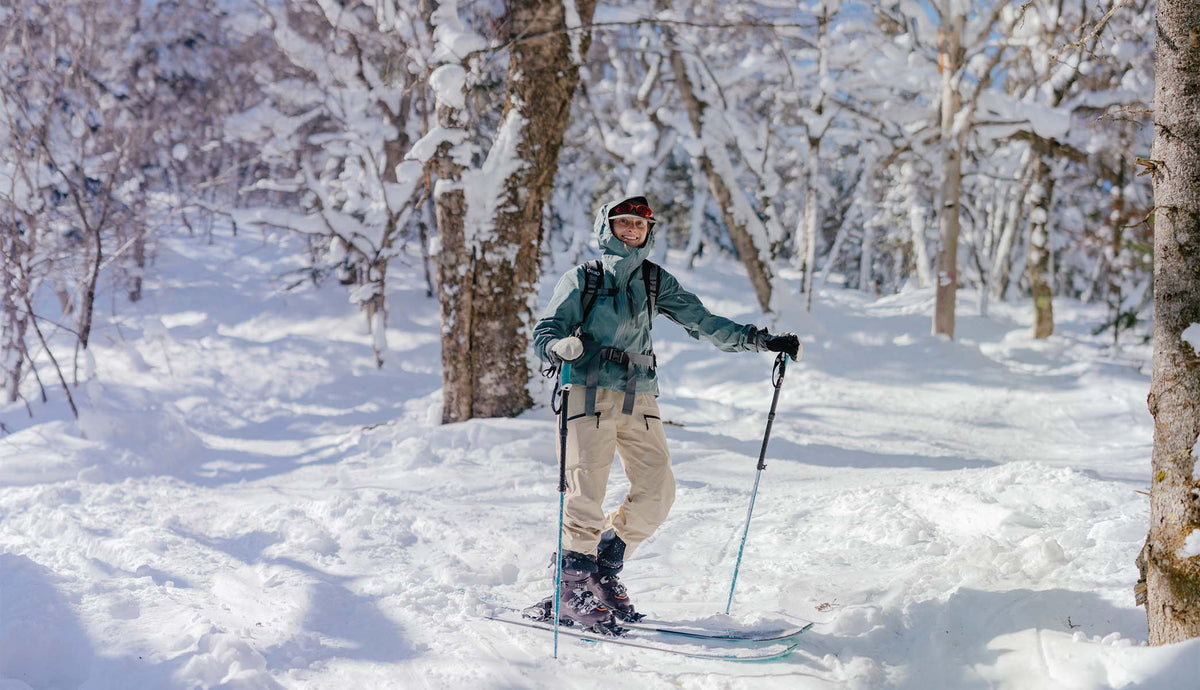 A skier stands in a snowy forest wearing a teal jacket, beige ski pants, and holding ski poles, with snow-covered trees in the background.