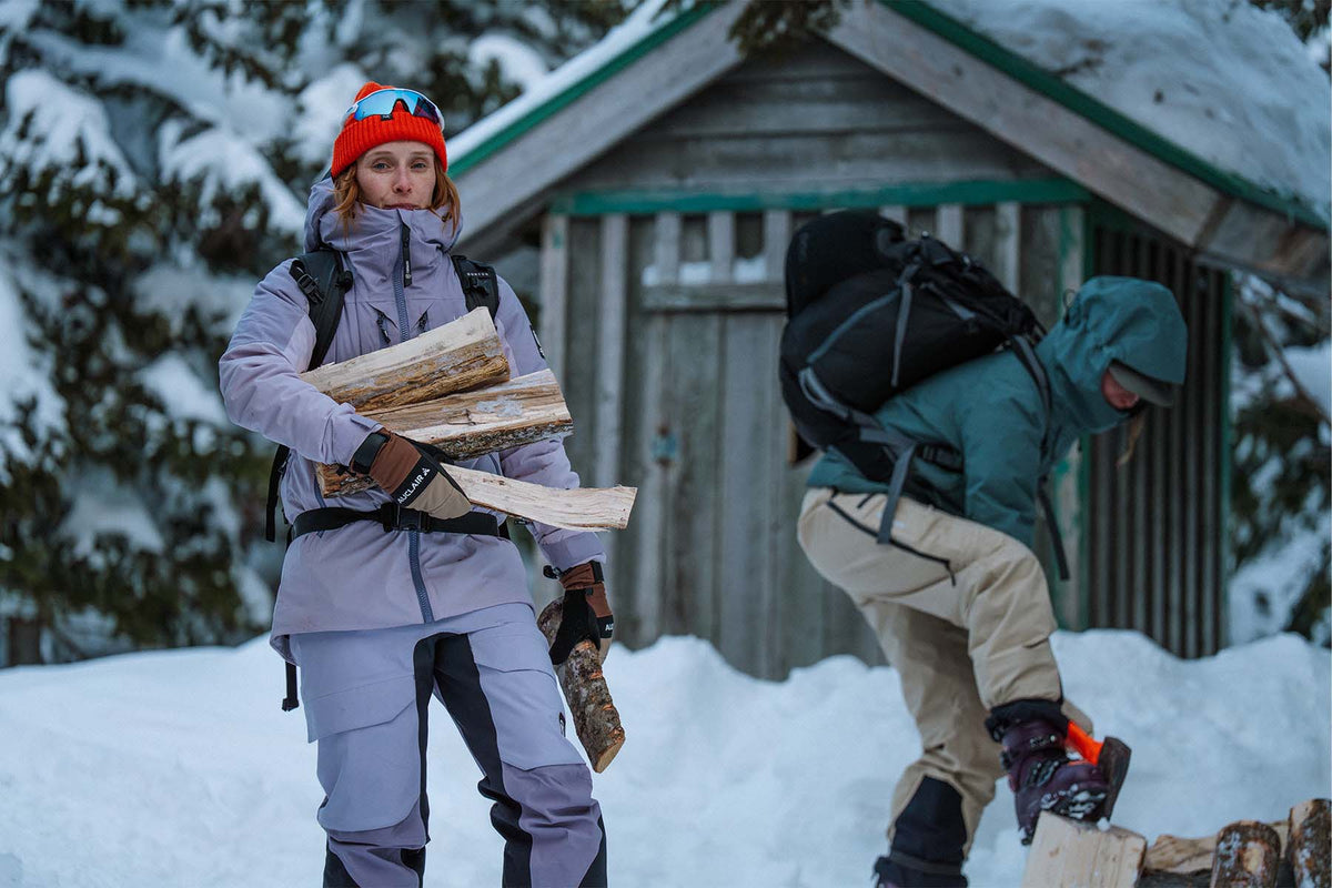 Two people in winter outdoor gear standing in deep snow in front of a small cabin. One person wears a light gray ski suit, a red beanie, and holds stacked firewood.