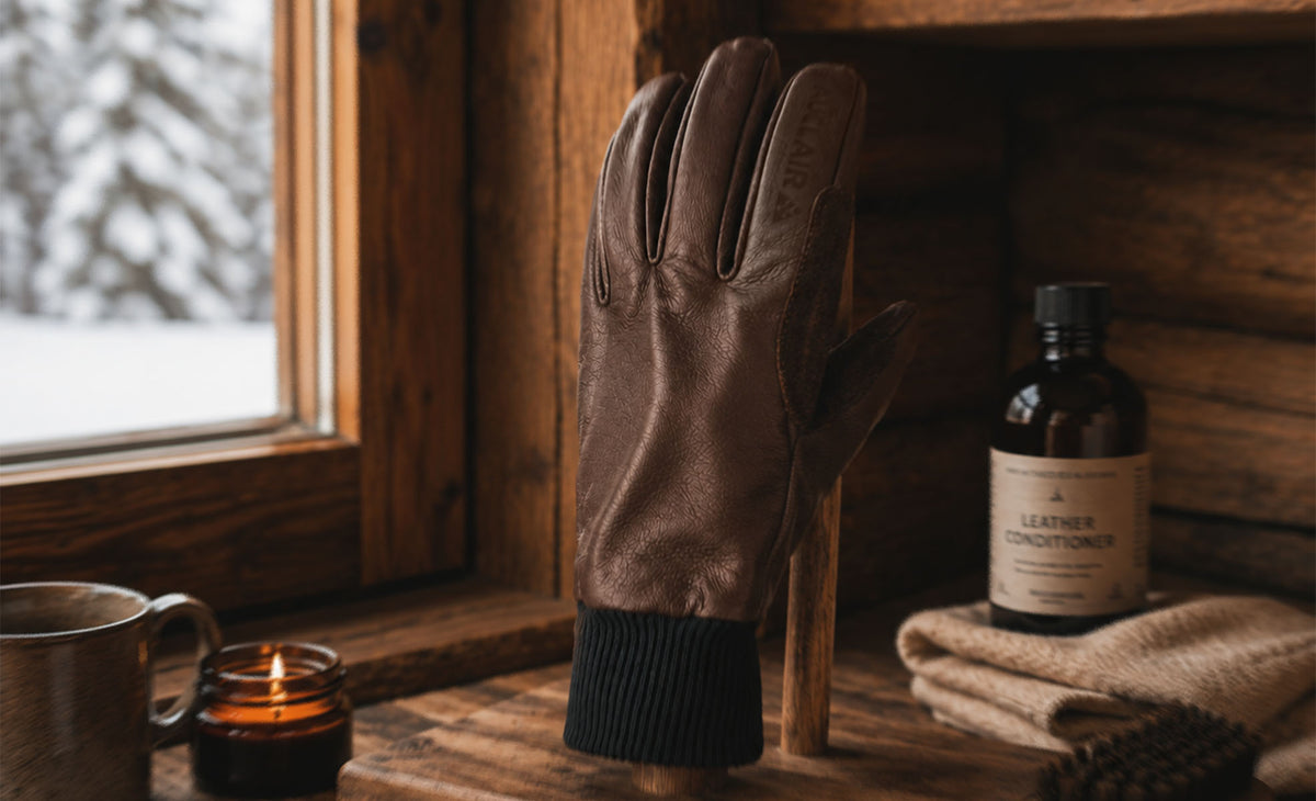 A brown leather glove displayed upright on a wooden stand on a rustic wooden table, with a snowy window, candle, mug, and leather conditioner bottle nearby.