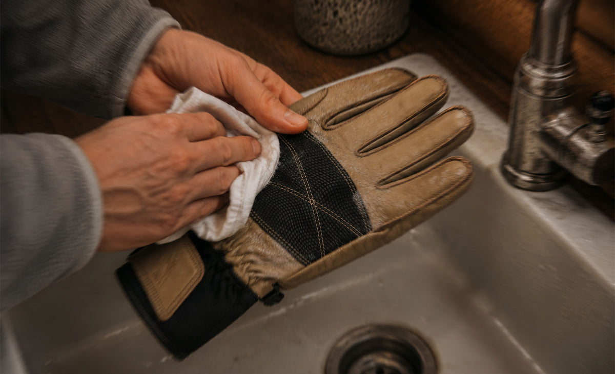 A close-up of a person's hands cleaning a tan and black work glove with a white cloth, held over a stainless steel kitchen sink.