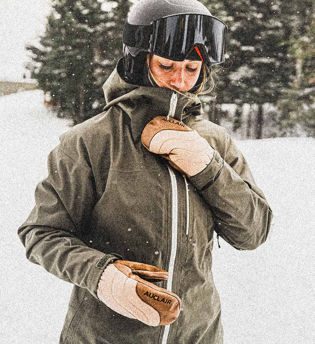 A person in a snowy outdoor setting wearing an olive green ski jacket, a dark helmet with goggles, and tan winter gloves with the brand name 'AUKLAIR' visible on the back of one glove.