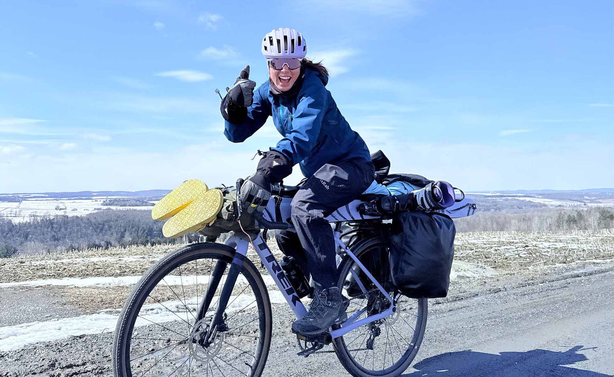 A woman wearing a white helmet and sunglasses rides a loaded bicycle on a road outdoors under a blue sky. She is dressed in a blue jacket and dark pants, with yellow shoes hanging from the handlebars and black panniers on the rear rack.