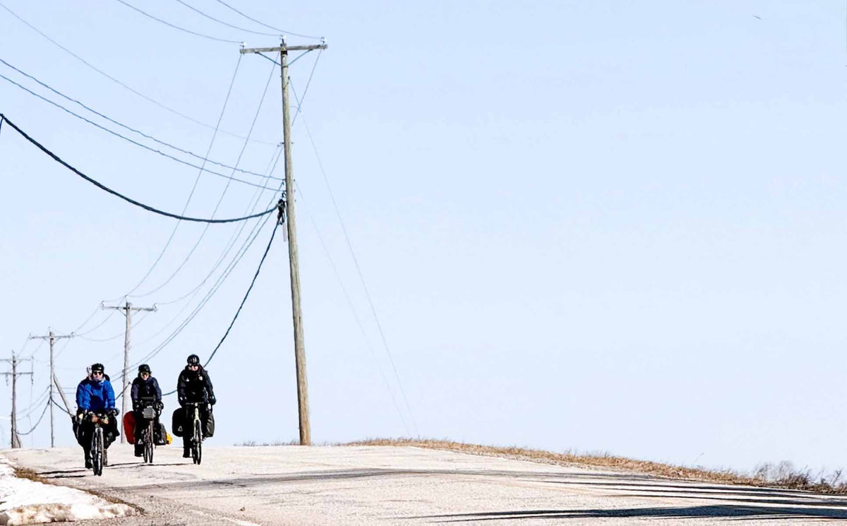 Three cyclists ride along a paved road under a clear blue sky, with utility poles and overhead wires lining the route.