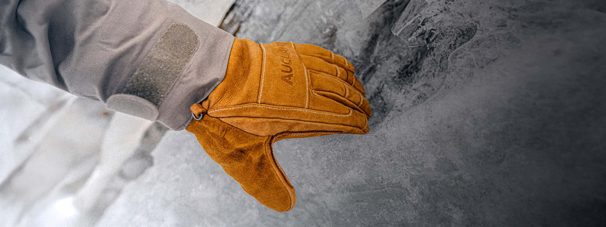 A close-up of a tan leather winter glove worn on a hand, with a gray sleeve visible and an icy outdoor background.