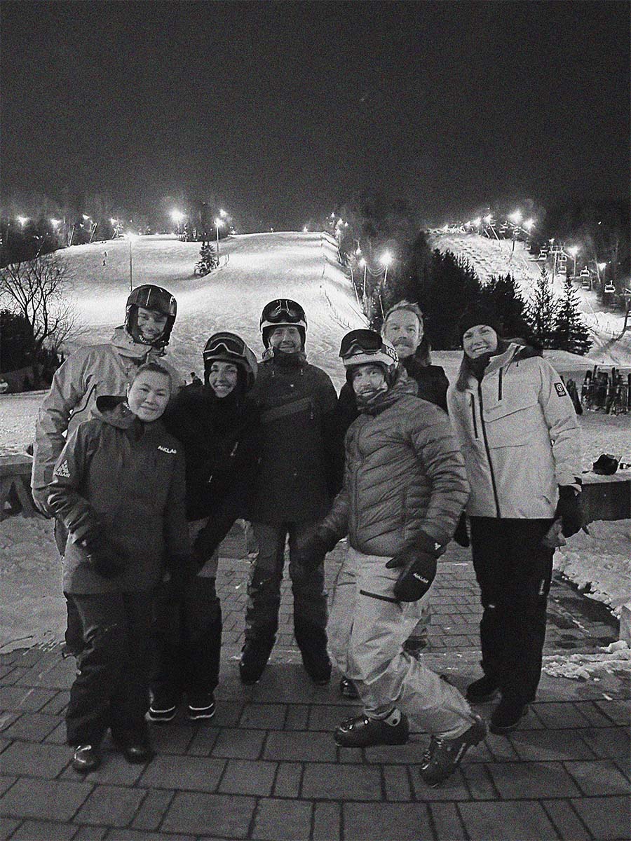 A black and white group photo of seven people wearing winter ski gear, helmets, and goggles, standing together on a paved area at night in front of a ski resort with snow-covered slopes and illuminated lights in the background.