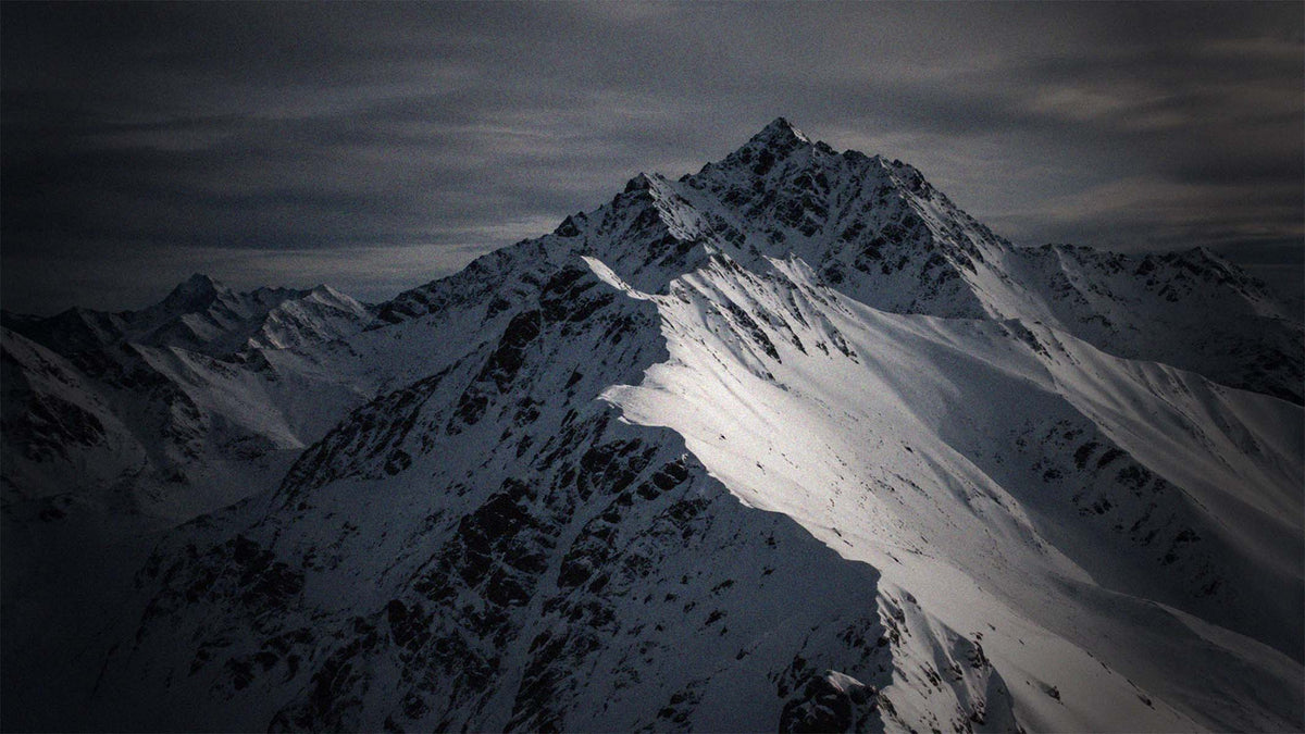 A dramatic mountain landscape with snow-covered peaks under a dark, cloudy sky. The image has high contrast with deep shadows and bright snow highlights.