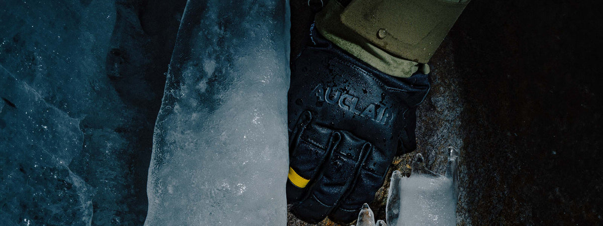 A close-up of a black tactical glove with 'AUGLAI' printed on the back, worn over a tan sleeve, positioned against a dark, icy background.